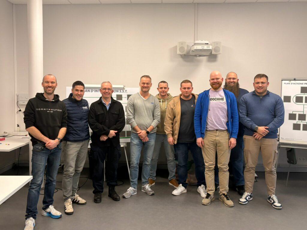 Photo de groupe de neuf hommes souriants posant dans une salle de formation devant des panneaux de 'Plan d'Intervention S.S.T. et de Plan d'Actions Prévention