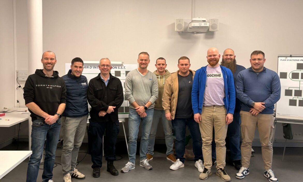 Photo de groupe de neuf hommes souriants posant dans une salle de formation devant des panneaux de 'Plan d'Intervention S.S.T. et de Plan d'Actions Prévention