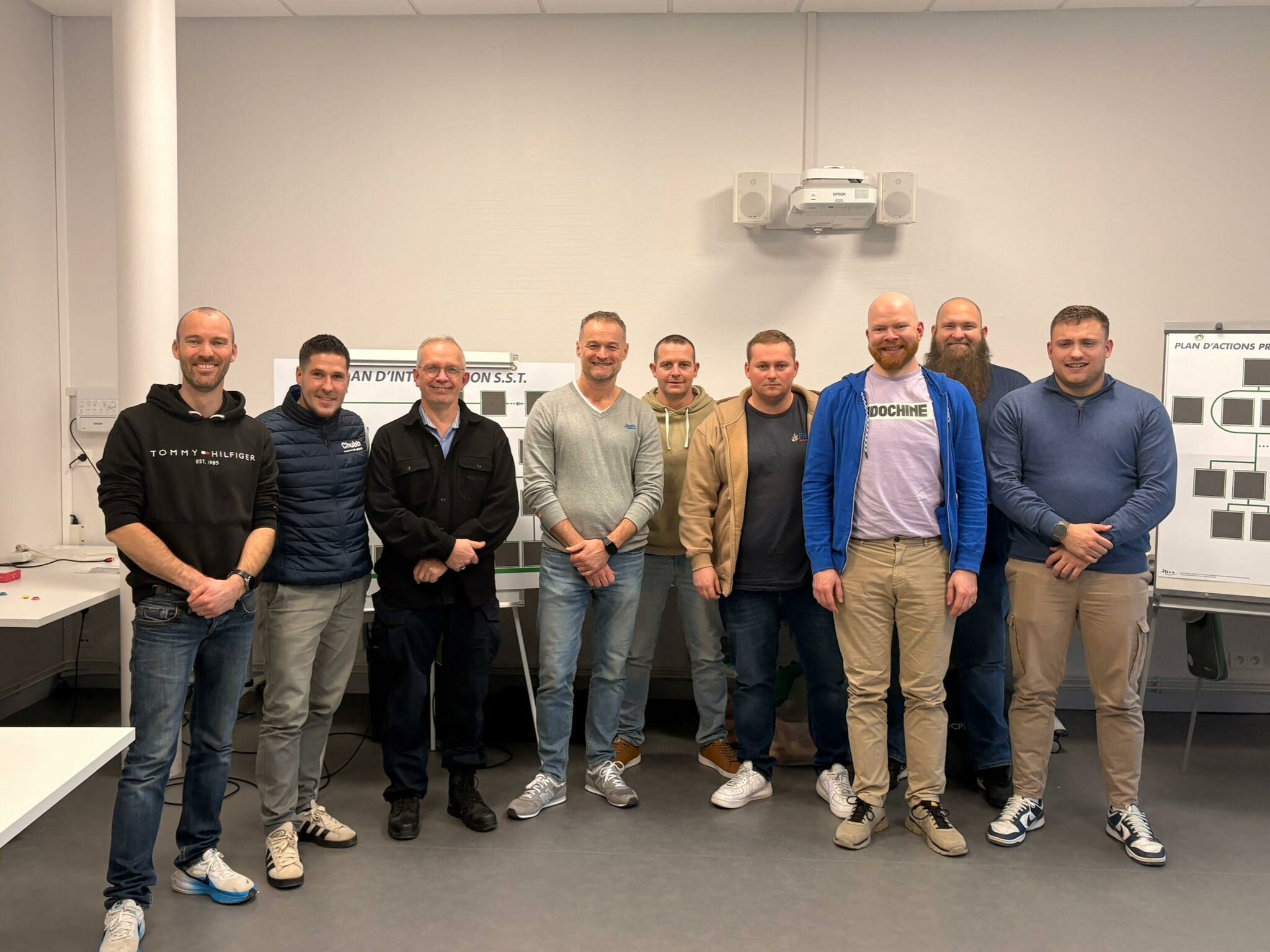 Photo de groupe de neuf hommes souriants posant dans une salle de formation devant des panneaux de 'Plan d'Intervention S.S.T. et de Plan d'Actions Prévention